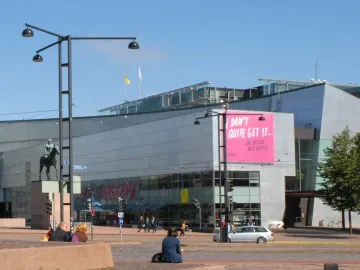 Marshal Mannerheim equestrian statue in front of a modern glass building in Helsinki, with a pedestrian in the foreground.