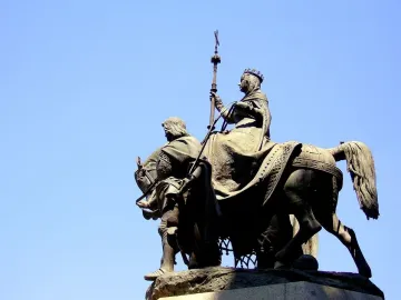 Monument to Isabella of Castile with a crowned woman on horseback and a bearded man beside the horse against a blue sky.