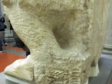 Lower section of the Palestrina Pietà in light marble, showing rough unfinished surfaces and chisel marks on a museum display platform.