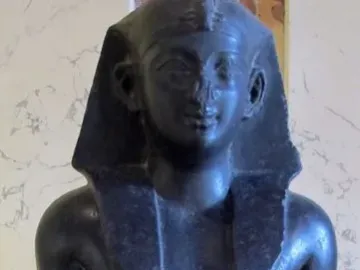 Upper torso of a Ptolemaic king statue in dark stone, with a nemes headdress and missing arms.