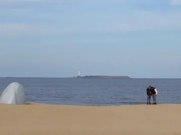 The Hand sculpture rising from the sand on a beach with the ocean and a distant island behind it.