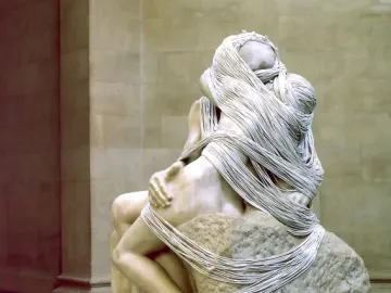 Marble sculpture of two figures embracing, draped with thin strings over a textured stone base.