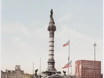 Soldiers\' and Sailors\' Monument rising from a stepped base in a landscaped park, with American flags nearby and city buildings in the background.