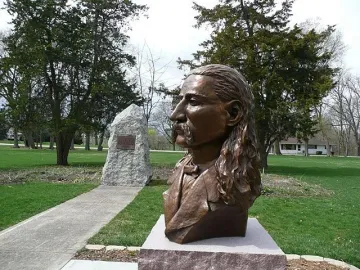 Bronze bust of Wild Bill Hickok on a textured pedestal with a pistol and hat relief, set behind a fence among pine trees.