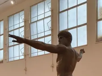 Artemision Bronze, a nude bronze male statue posed with one arm extended and the other drawn back in a museum display.