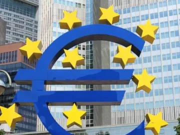 Blue euro sculpture with yellow stars in front of modern glass buildings, with people gathered below it.