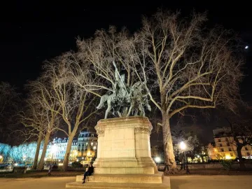 Illuminated bronze monument of Charlemagne and His Leudes with a rider and two standing figures on a tall stone pedestal at night.