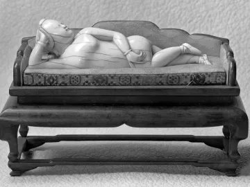Doctor\'s Lady, a light-colored female figure reclining on a small couch with a patterned cushion, shown on a wooden stand.