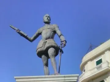 Bronze statue of John of Austria in historical armor on a tall stone pedestal, with one arm extended and a sword in the other hand.