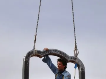 A large dark metal frame for Memoria Urbana hangs from chains while a worker crouches inside it with a wrench.