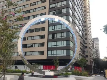 The Eye of Mexico sculpture in an urban plaza beside a tall gridded building, framed by trees and city streets.