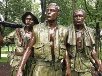 The Three Soldiers bronze statue with three wet-looking soldiers standing outdoors among trees.