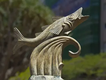 The Chicharro Sculpture shows a metallic fish leaping from a stylized wave outdoors.