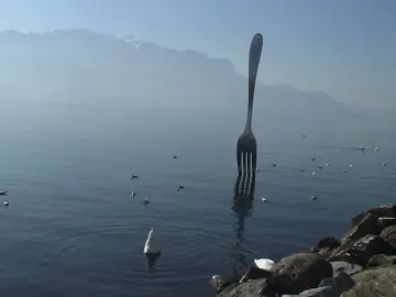 The Fork of Vevey sculpture rises from the water with mountains and birds in the background.
