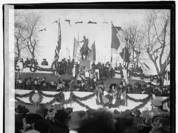 Joan of Arc equestrian statue on a pedestal at an outdoor gathering, with flags, garlands, spectators, and bare trees around it.