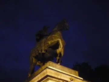 Illuminated bronze equestrian monument to Peter I on a tall stone pedestal with a double-headed eagle and Cyrillic inscriptions.