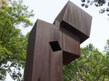 El Llarg Viatge, a tall abstract reddish-brown metal sculpture with a square opening, standing in an urban park.
