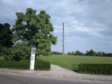 The Endless Column standing in a grassy park beside a paved road, with a tree partially obscuring the left side.
