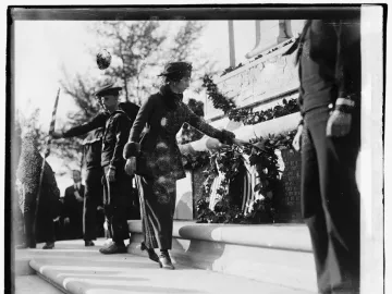 A woman places a wreath on the John Paul Jones Memorial while men in naval uniforms stand nearby on the steps.