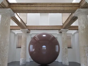 Large Red Sphere displayed indoors between weathered classical pillars.