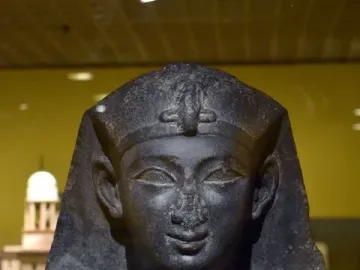 Upper torso and head of a dark basalt statue of a Ptolemaic king, with a damaged nose and traditional headdress.