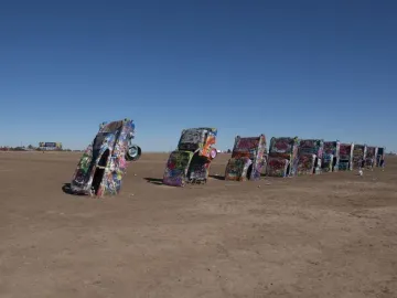 Cadillac Ranch cars covered in graffiti stand buried nose-first in a flat dirt field under a clear blue sky.