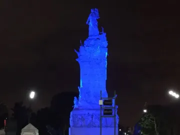 The Monument to the Carta Magna and Four Regions of Argentina lit in vivid blue at night, with a small stone pillar, streetlights, and trees nearby.