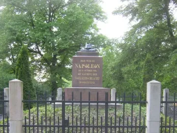 Napoleonstein stone monument behind a dark metal fence and stone pillars, surrounded by trees and greenery.