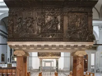 The Passion Pulpit in a church interior, with dark bronze relief carvings on red marble columns above a checkered floor and wooden pews.