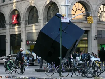 The Alamo sculpture, a large dark tilted cube, stands in an urban street scene near a stone building and street signs.