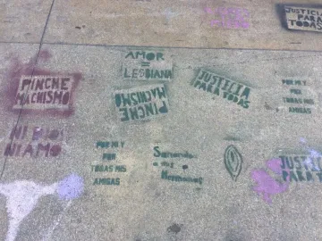 Concrete sidewalk covered with green and purple stencil graffiti in Spanish, including feminist slogans and symbols, near the Antimonumenta.