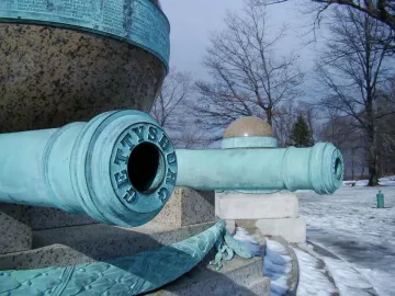 Close-up of two bronze cannons at the Battle Monument, with GETTYSBURG visible on the front cannon and a snow-dusted monument behind them.