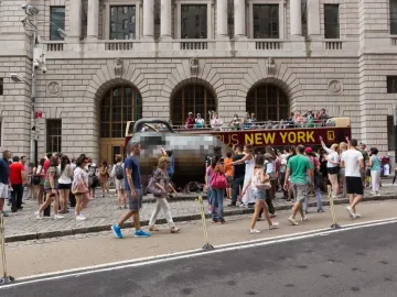 Charging Bull sculpture on a city street surrounded by pedestrians, with a tour bus and stone building in the background.