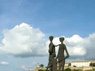 Monument to the Victims of Fascism with bronze figures on a brick platform beside flowering bushes and a river.