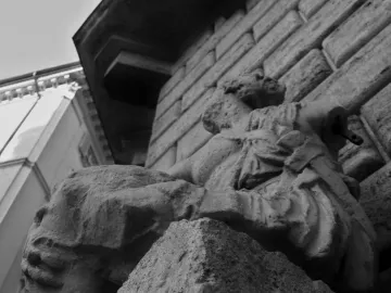 Weathered Pasquino stone statue with eroded draped figures beside a stone building and a metal rod protruding from the shoulder.