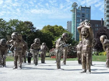 A-maze-ing Laughter sculpture with metallic human-like figures in an outdoor plaza, with buildings and trees behind it.