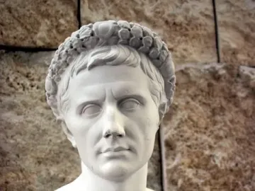 Bust of Augustus wearing a gemmed crown, displayed on a blue stand against a stone wall.