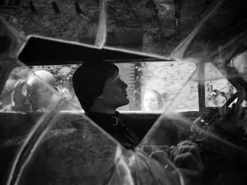 Surreal black and white scene of Rainy Taxi seen through a fractured windshield, with a mannequin in the driver\'s seat and reflections of two people in the windows.