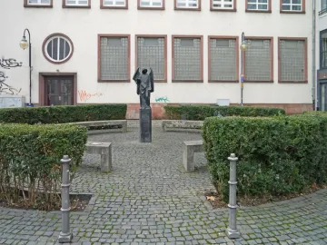 The Frankfurter Engel statue stands on a pedestal in a paved plaza with hedges and benches in front of a building with a circular window.