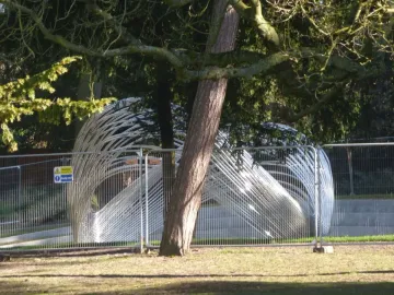 Infinite Wave sculpture behind a temporary metal fence in a wooded park setting.