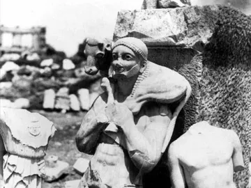 A headless nude marble torso, identified as the Kritios Boy, standing among fragmented ancient statues and ruins.