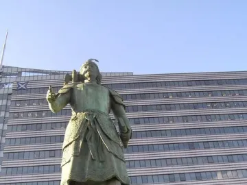 Marco Polo statue in historical attire with a bird on his arm, standing on a stone pedestal.