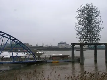 Quantum Cloud sculpture rising above the River Thames near a blue arched bridge and waterfront buildings.