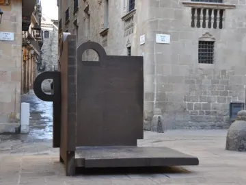 Topos V, a dark geometric sculpture by Eduardo Chillida, stands in a stone-paved courtyard beside old stone buildings.