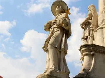 Weathered stone figure from the Holy Trinity Monument holding a golden cross and looking upward, with another robed statue partly visible beside it.