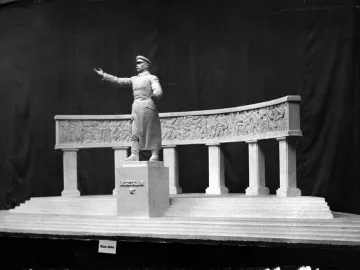 A model for a Stalin monument showing a figure in a long coat and cap before a curved colonnade with relief sculptures.