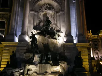 The Fountain of King Matthias illuminated at night, with dark statues on tiered rocky levels and a central figure above.
