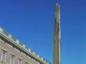 The Obelisk at Slottsbacken stands in a paved square beside a long building under a clear blue sky.