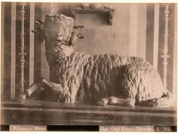 Resting bronze ram with curled horns on a long pedestal, shown against a striped and pillared wall backdrop.