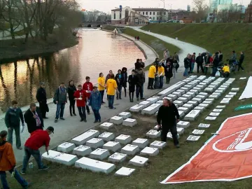 People gathered around the Keyboard Monument, with one person painting a key on the large concrete keyboard beside a river.